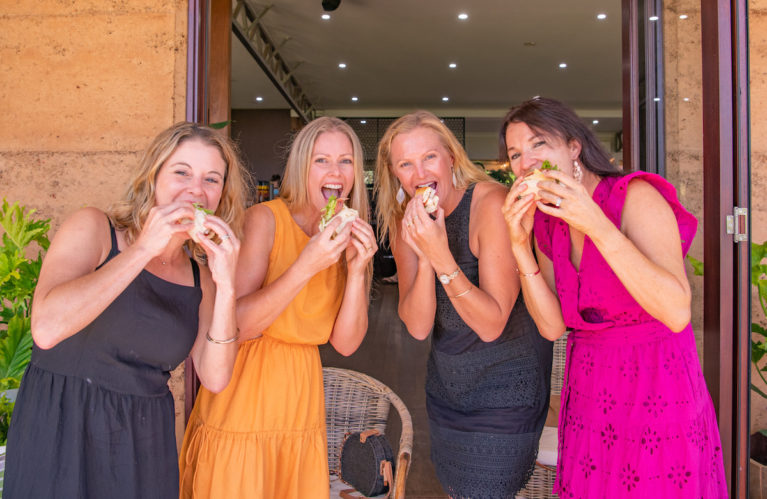 4 ladies in dresses smiling and eating a roll at a winery